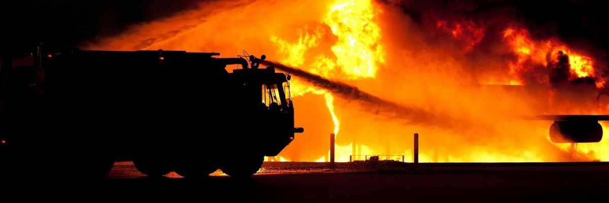 Das Bild zeigt einen Großbrand der von einem Feuerwehrfahrzeug gelöscht wird. Es ist nur die Silhouette des Feuerwehrfahrzeuges zusehen. 