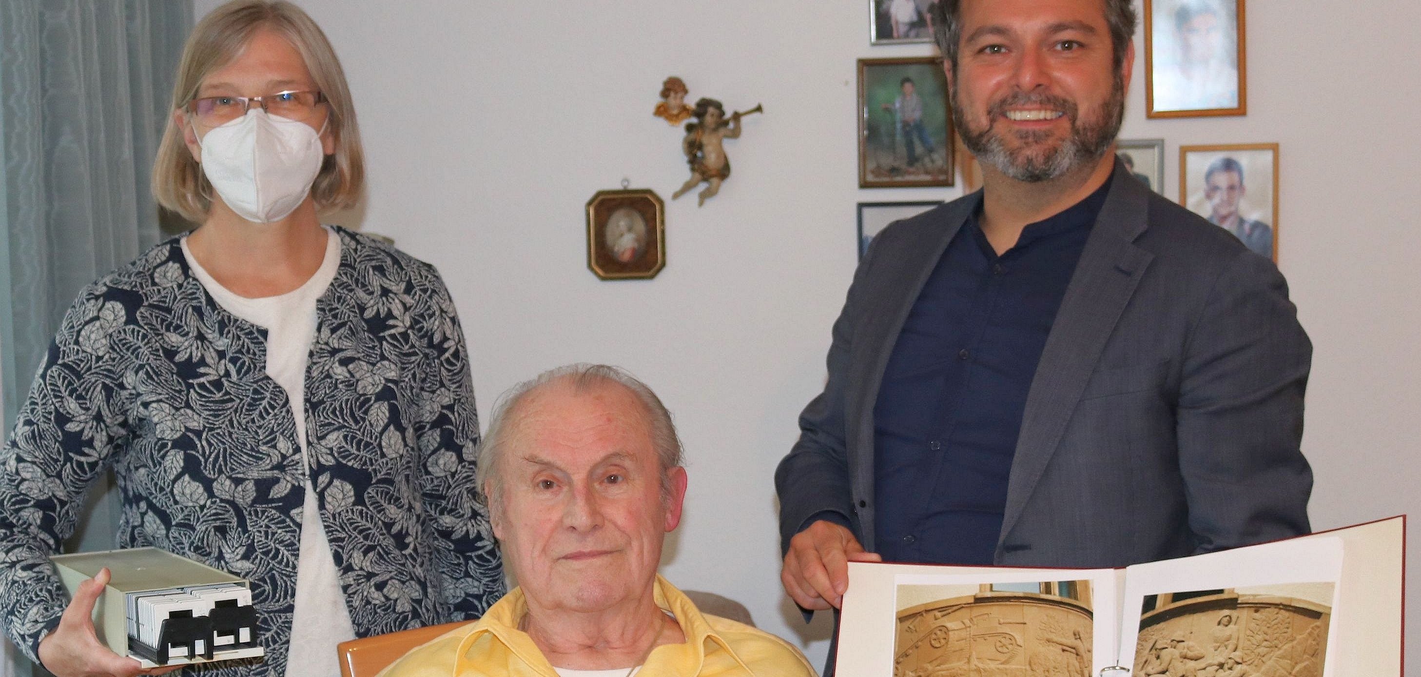 Walter Kastner, der in der Mitte des Fotos auf einem Stuhl sitz, Anette Vinnen, Leiterin des Stadtarchivs, die links steht und Bürgermeister Daniel Christian Glöckner. halten Fotos von Walter Kastner in die Kamera.