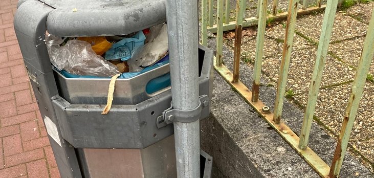 Ein mit allerlei Müll vollgestopfter öffentlicher Abfalleimer. Daneben steht eine Plastiktüte mit Glasflaschen. 