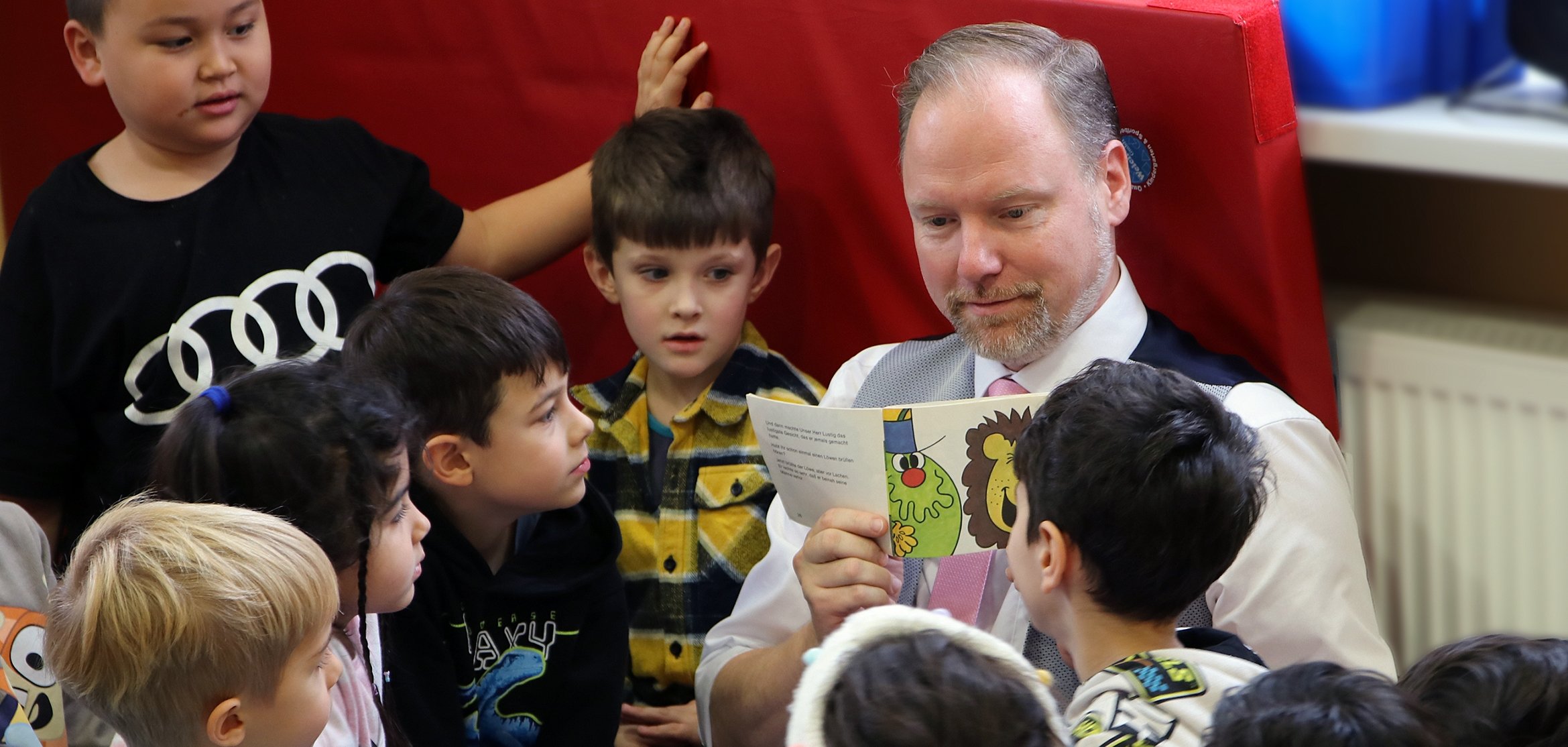 Der Bürgermeister sitzt auf einer Turnmatte, umringt von einer Gruppe von Kindern.