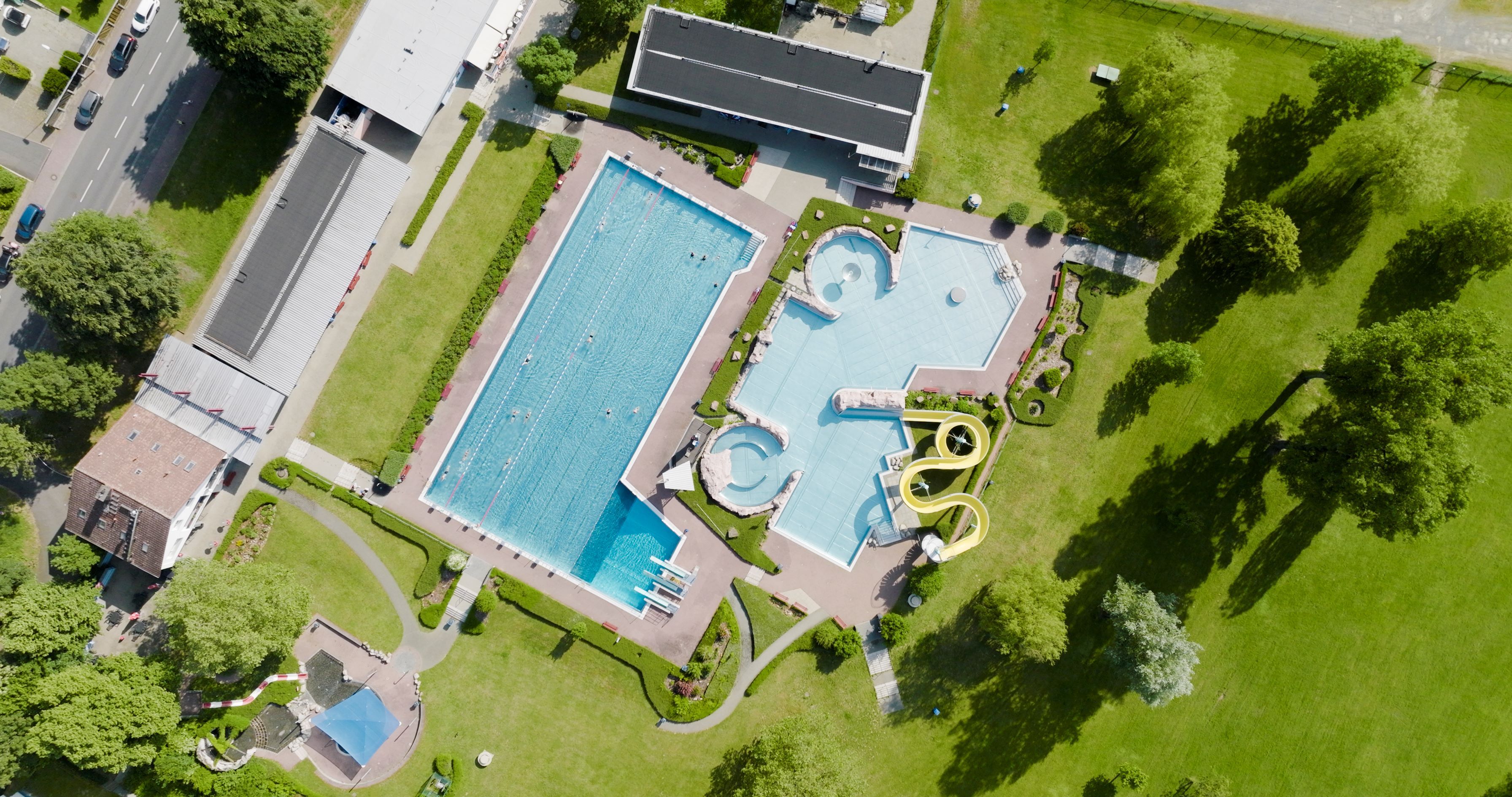 Luftaufnahme des Freibads mit Gebäuden, Becken und Liegewiese aus dem Sommer.