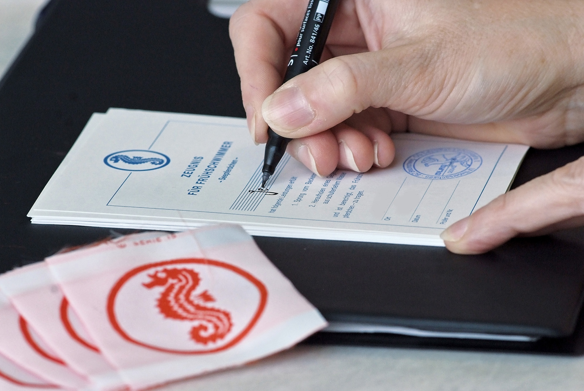Eine menschliche Hand beim Ausfüllen einer DRLG Urkunde. Im Vordergrund liegen Seepferdchenaufnäher. 