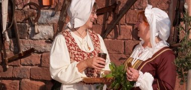 Zwei lachende Frauen in historischen Gewändern vor einer Sandsteinmauer.