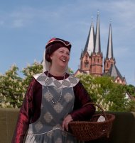 Eine lachende Frau im mittelalterlichen weinroten Kostüm aus dem Jahr 1389 mit einem geflochtenen Korb am Arm. Im Hintergrund die Türme der Marienkirche.