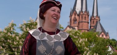Eine lachende Frau im mittelalterlichen weinroten Kostüm aus dem Jahr 1389 mit einem geflochtenen Korb am Arm. Im Hintergrund die Türme der Marienkirche.