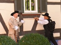 Holzauge sei wachsam Mann im Kostüm eines Zimmermanns aus dem 18. Jht und eine Frau in Zimmermannskleidung der heutigen Zeit vor der Fassade eines Fachwerkhauses.