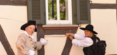 Mann im Kostüm eines Zimmermanns aus dem 18. Jht und eine Frau in Zimmermannskleidung der heutigen Zeit vor der Fassade eines Fachwerkhauses.