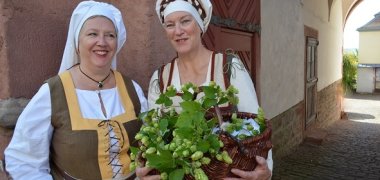 2 Frauen in Kostümen aus dem 16. Jht. Die rechte trägt einen geflochtenen Korb mit Hopfenblüten. Sie stehen vor dem Tor zu einem Innenhof.
