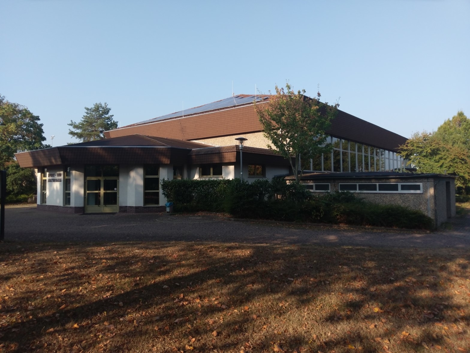 Man sieht ein Gebäude mit einem Achteckigen Anbau. Das Gebäude hat eine Große Fensterfront und es wachsen einige Bäume im Hof des Gebäudes.  