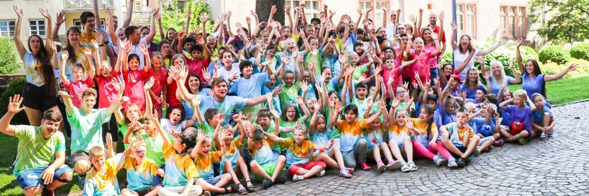 Gruppenbild vom Kindersommer. Alle haben bunte T-Shirts an und ihre Arme nach oben gestreckt.