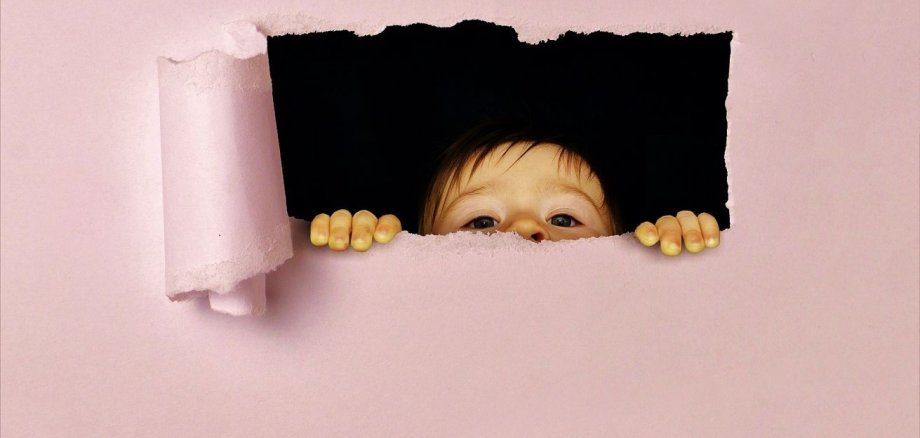 Ein Kind lugt durch ein rechteckiges Loch hinter einer Papierwand hervor. 