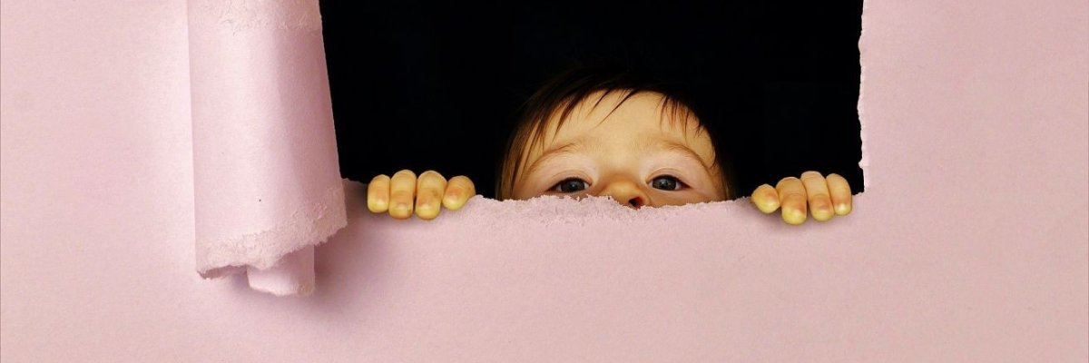 Ein Kind lugt durch ein rechteckiges Loch hinter einer Papierwand hervor. 