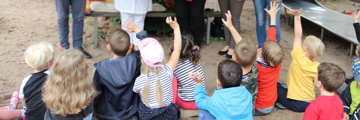 Kinder sitzen auf dem Sandboden der Kita Rappelkiste. Sie sind nur von hinten zu sehen.