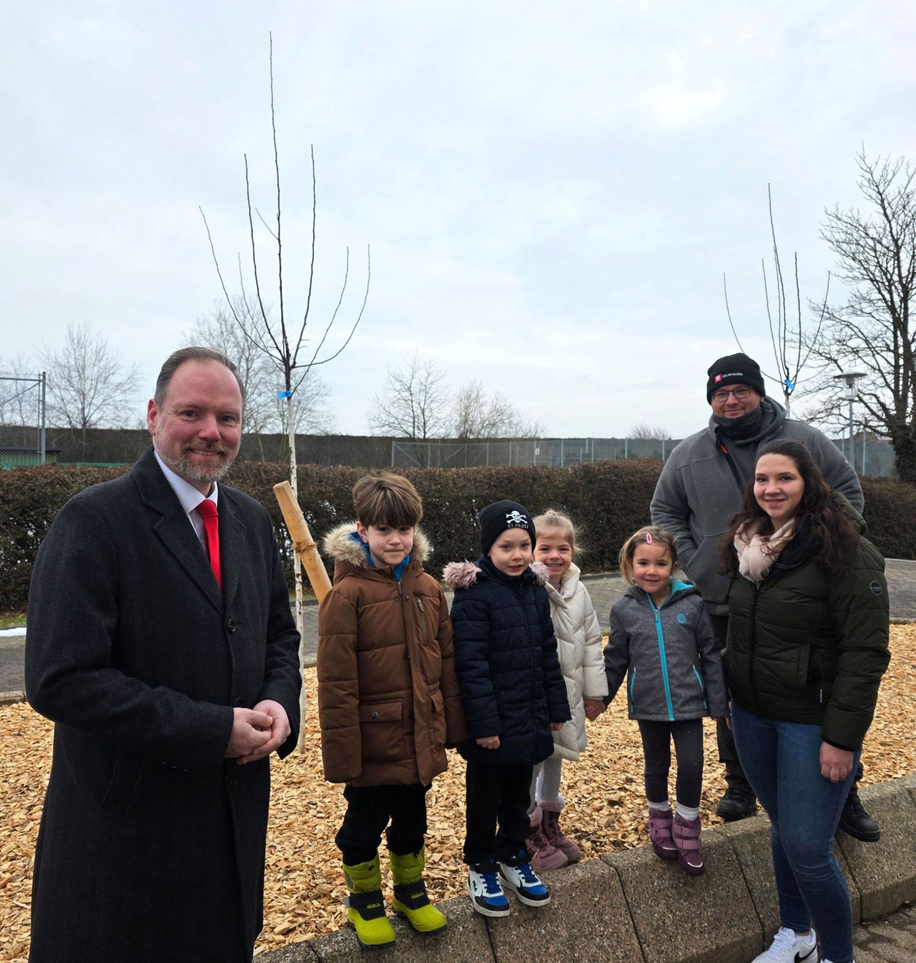 Drei Erwachsene und vier Kinder stehen gemeinsam an den zwei neu gepflanzten Apfelbäumen an der Sport- und Kulturhalle in Meerholz. 
