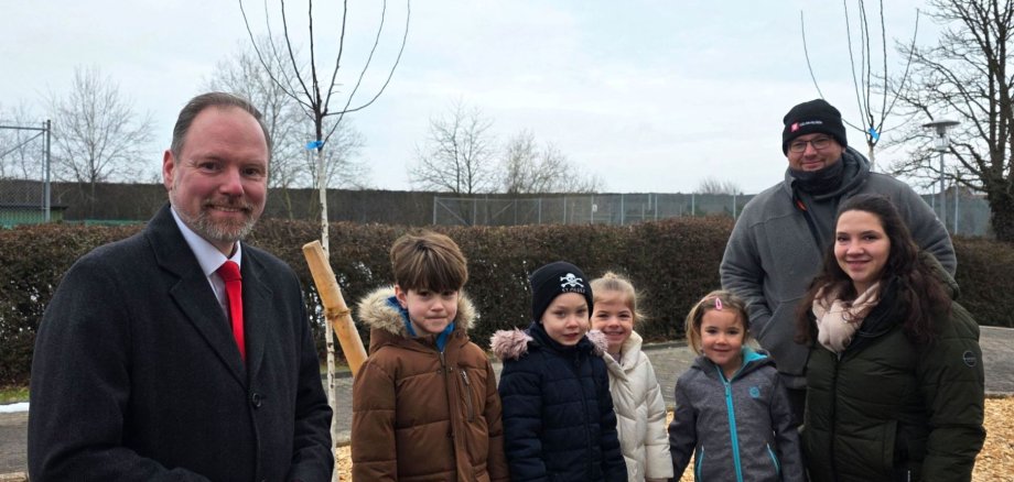 Drei Erwachsene und vier Kinder stehen gemeinsam an den zwei neu gepflanzten Apfelbäumen an der Sport- und Kulturhalle in Meerholz. 