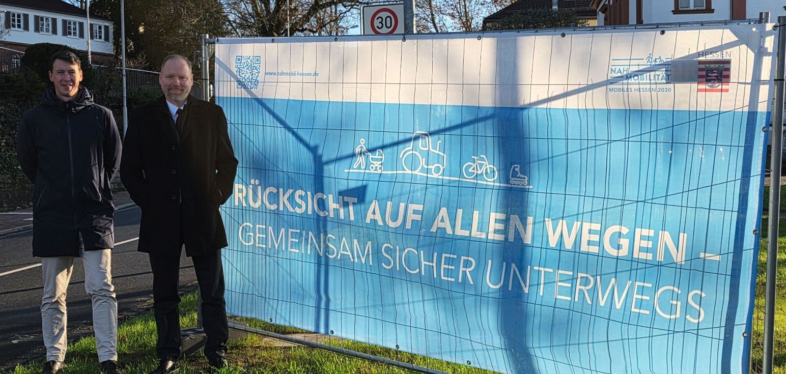 Zwei Männer stehen auf einer Wiese neben einem Banner, auf dem zur Verkehrssicherheit aufgerufen wird.