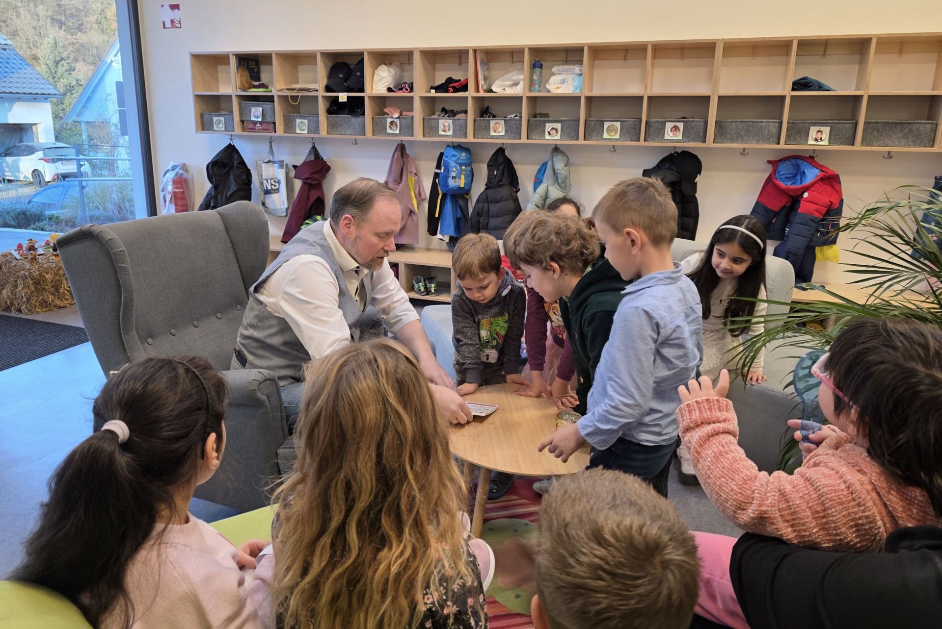 Der Bürgermeister sitzt auf einem Sessel und liest aus einem Buch. Kinder stehen und sitzen um ihn herum und hören zu. 