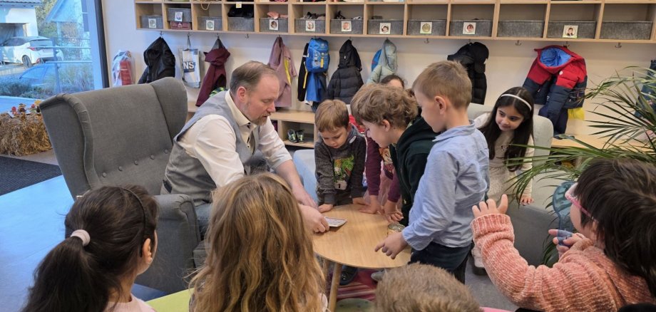 Bundesweiter Vorlesetag in Gelnhausen Der Bürgermeister sitzt auf einem Sessel und liest aus einem Buch. Kinder stehen und sitzen um ihn herum und hören zu.