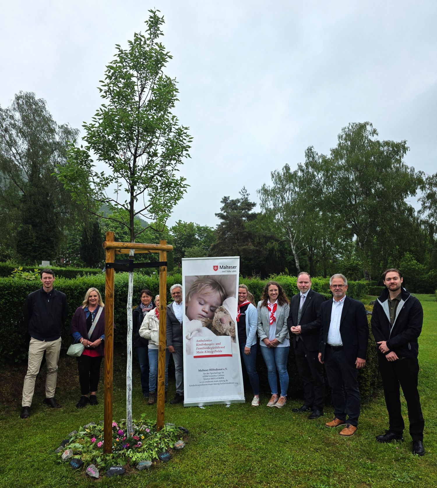 Bürgermeister Christian Litzinger und Vertreter der Malteser stehen mit weiteren Teilnehmenden an einem neu gepflanzten Gedenkbaum auf dem Friedhof in Gelnhausen-Mitte und weihen den neuen Gedenkort ein.