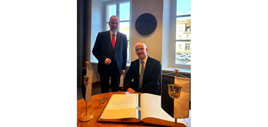 Pfarrer Rainer Schomburg sitz im Sitzungssaal des Rathauses vor einem Tisch auf dem das Goldene Buch der Stadt Gelnhausen liegt. Bürgermeister Christian Litzinger steht neben ihm. 