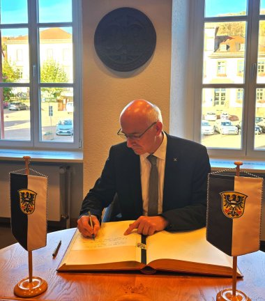 Pfarrer Rainer Schomburg sitz im Sitzungssaal des Rathauses auf einem Stuhl und trägt sich im Goldenen Buch der Stadt Gelnhausen ein, das vor ihm auf dem Tisch liegt. 