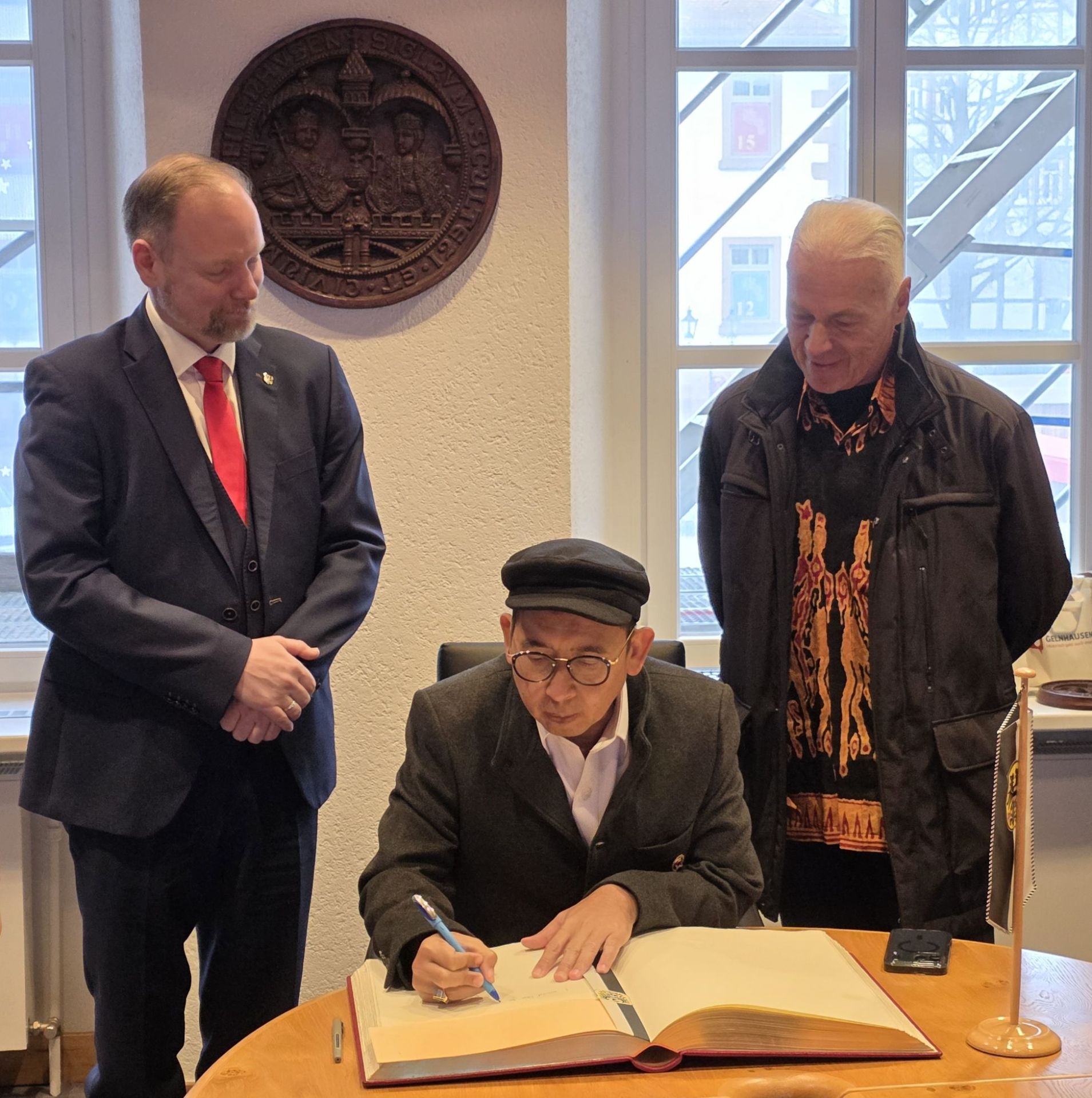 Ein Mann sitzt an einem Tisch und schreibt in ein Buch. Zwei andere Männer stehen daneben und sehen ihm zu. 