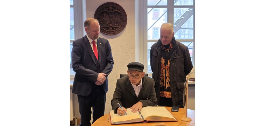 Ein Mann sitzt an einem Tisch und schreibt in ein Buch. Zwei andere Männer stehen daneben und sehen ihm zu. 