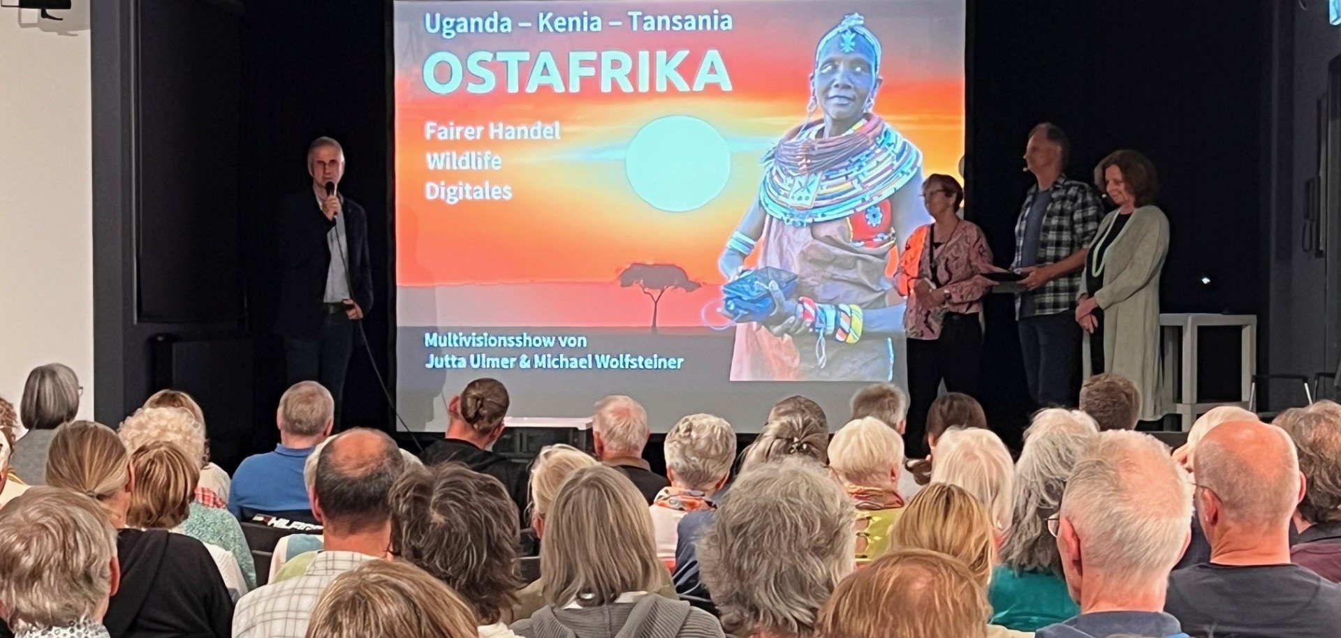 Viele Menschen sitzen in der Kulturherberge in Gelnhausen für einen Vortrag über Ostafrika und schauen auf die Bühne, auf der mehrere Menschen stehen. 
