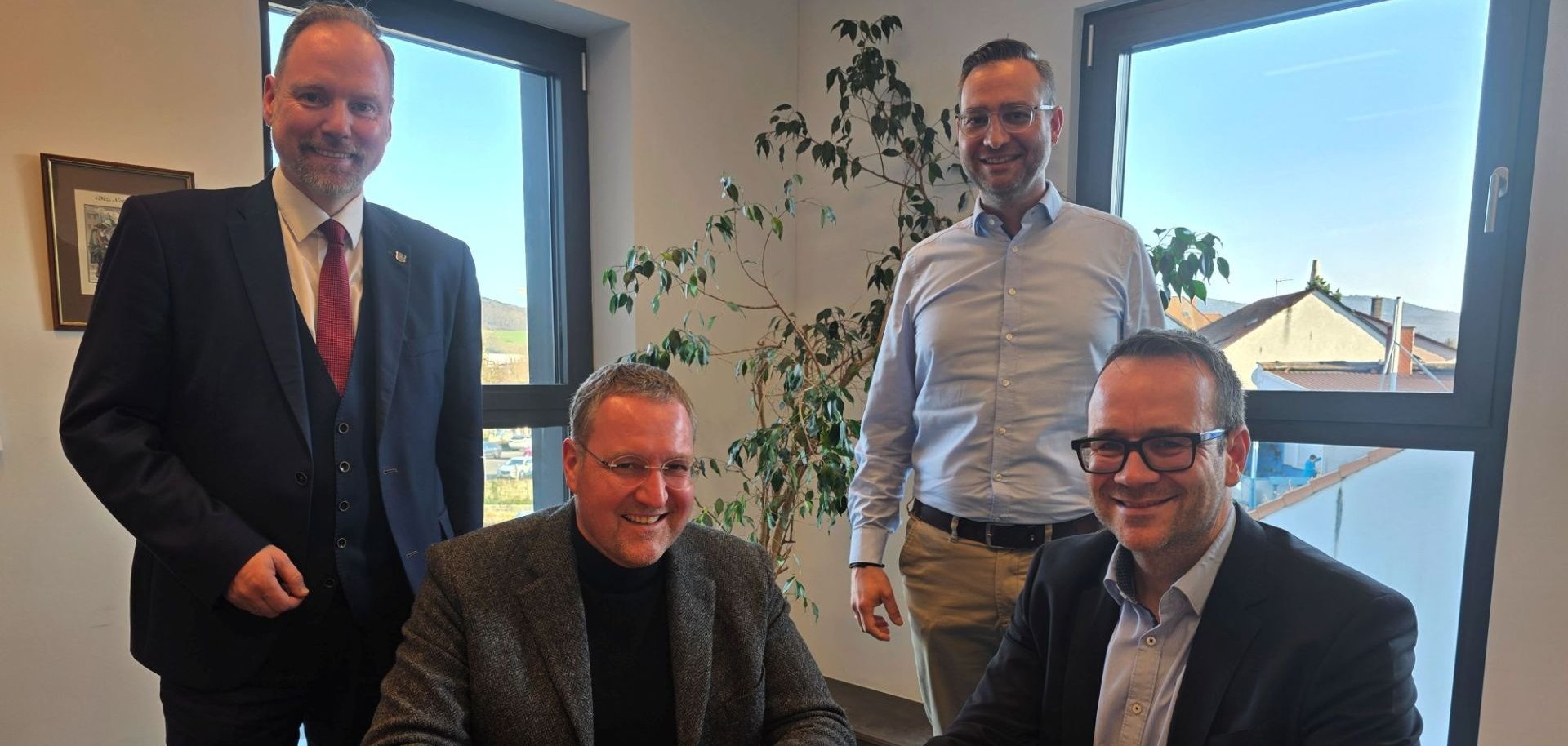 Zwei Personen sitzen in einem Raum an einem Tisch auf dem ein Dokument zur Unterzeichnung liegt. Zwei Personen stehen hinter ihnen. 