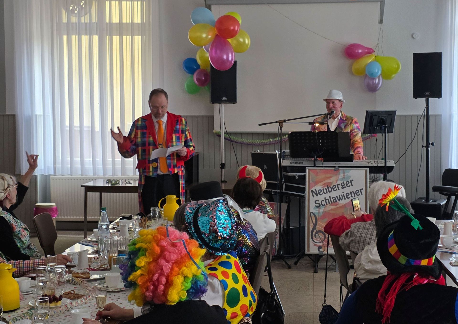Der Bürgermeister steht in einem Raum verkleidet in buntem Sacko und erzählt eine Geschichte. Im Saal sitzen verkleidete Seniorinnen und Senioren und hören zu. 