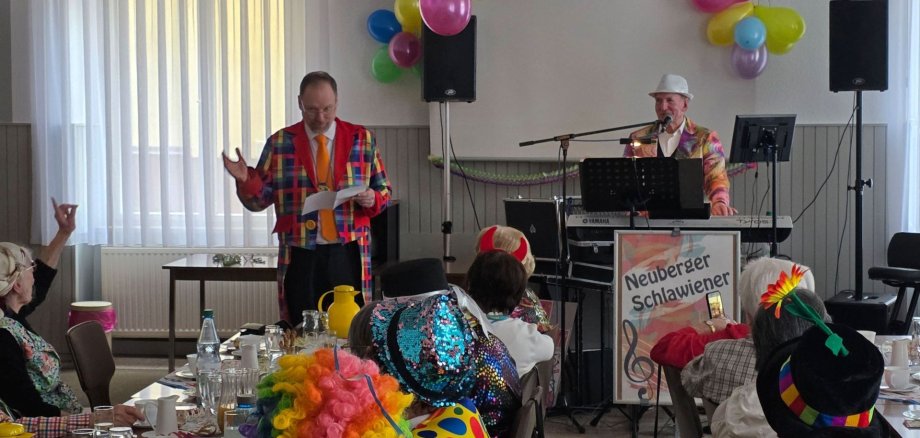 Seniorenclubs feiern Fasching in Gelnhausen Der Bürgermeister steht in einem Raum verkleidet in buntem Sacko und erzählt eine Geschichte. Im Saal sitzen verkleidete Seniorinnen und Senioren und hören zu.