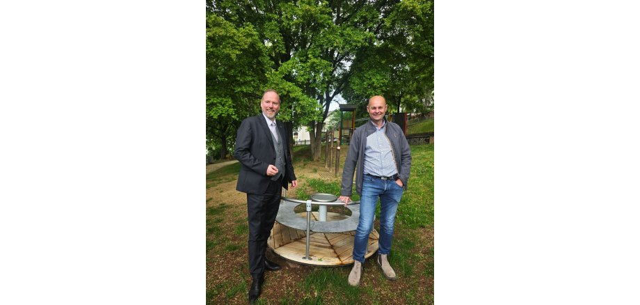 Bürgermeister Christian Litzinger steht links und Projektleiter Thorsten Amend rechts auf dem Röther Spielplatz „Im Goldberg“ an einem neuen Karussell.    