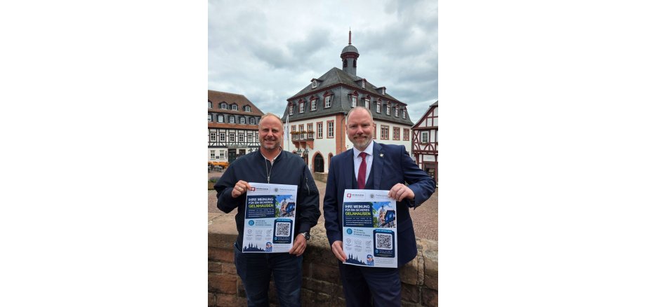 KOMPASS-Kurzbefragung zur Sicherheit in Gelnhausen Bürgermeister Christian Litzinger (rechts) und Ordnungsamtsleiter Bastian Metzler (links) stehen auf dem Obermarkt und halten jeweils ein Plakat in die Kamera, das zur Teilnahme an der KOMPASS-Kurzbefragung aufruft.