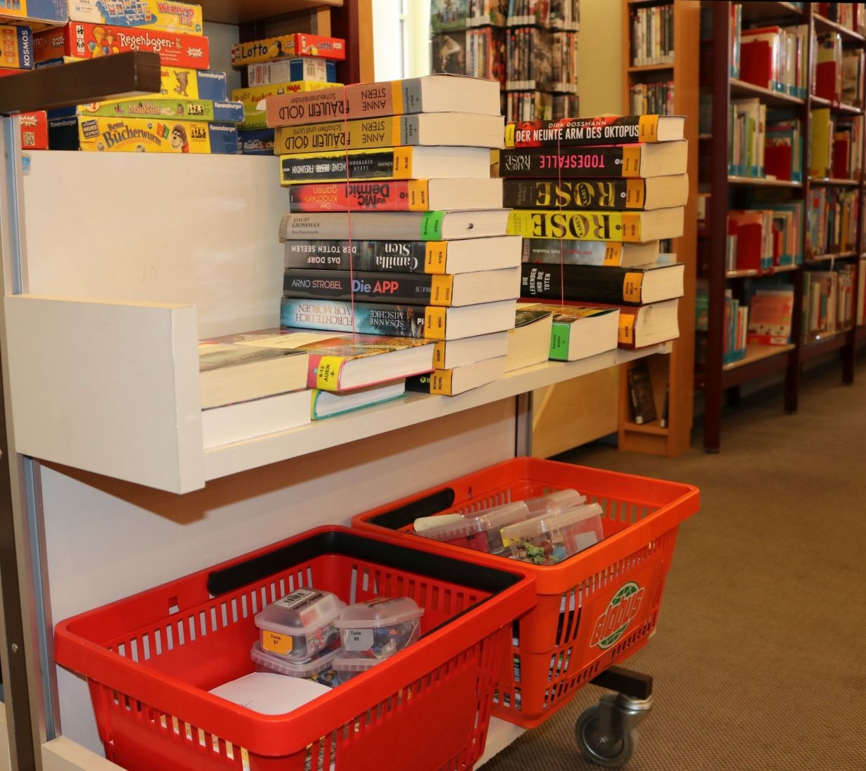 Abholservice Stadtbücherei Auf einem Rollwagen sind auf der oberen Ebene Bücher gestapelt, auf der unteren Ebene orangefarbene Körbe mit Medien wie Tonies, Playstationspiele etc. Im Hintergrund ist ein Teil der Bücherei mit gut bestückten Regalen zu sehen.