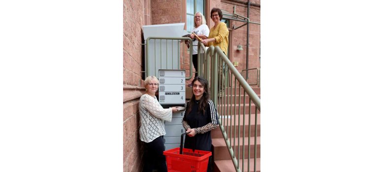 Graue Abholstation mit Fächern im Vordergrund, mit Zwei Frauen, die diese mit Büchern befüllen. Am oberen Ende der Treppe zwei weitere Frauen, die Medien in die Rückgabebox legen.