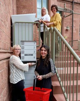 Abholstation Graue Abholstation mit Fächern im Vordergrund, mit Zwei Frauen, die diese mit Büchern befüllen. Am oberen Ende der Treppe zwei weitere Frauen, die Medien in die Rückgabebox legen.