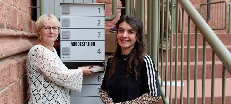 Abholstation Graue Abholstation mit Fächern im Vordergrund, mit Zwei Frauen, die diese mit Büchern befüllen. Am oberen Ende der Treppe zwei weitere Frauen, die Medien in die Rückgabebox legen.