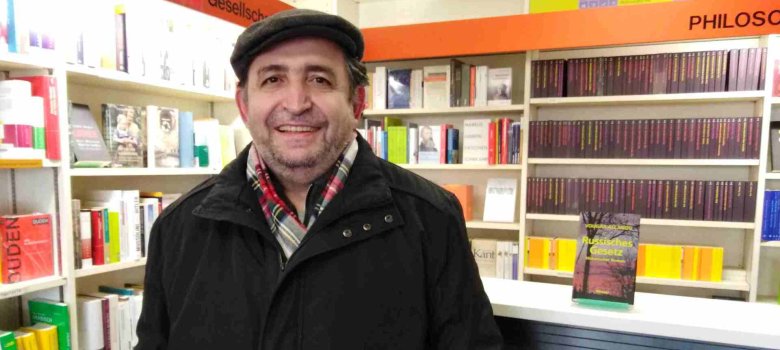 Autor mit schwarzer Jacke und Hut stehend in einer Buchhandlung mit Regalen voller Bücher im Hintergrund