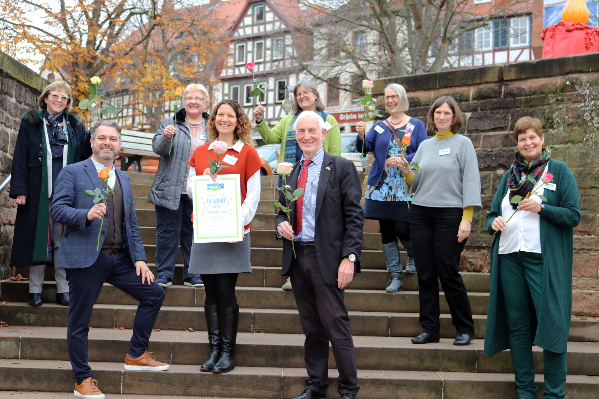 Das Bild zeigt Bürgermeister Daniel Chr. Glöckner, Bixi Scheffler, Leiterin der Steuerungsgruppe, den Fairtrade-Ehrenbotschafter Manfred Holz und Mitglieder der Steuerungsgruppe auf der Treppe zum Brunnen am Obermarkt. Alle lachen und halten jeweils eine fair gehandelte Rose in die Höhe.