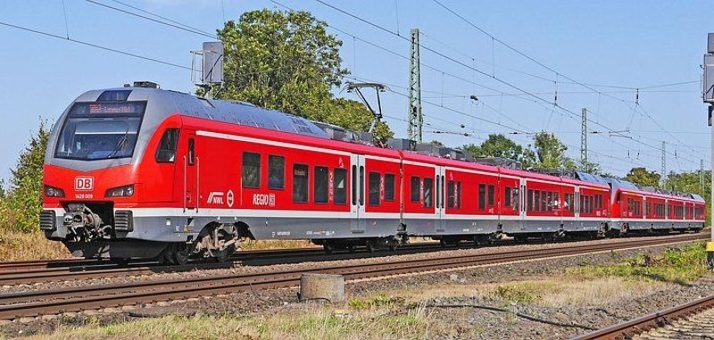Bild einer roten Bahn auf Gleisen im Hintergrund sieht man Bäume. 
