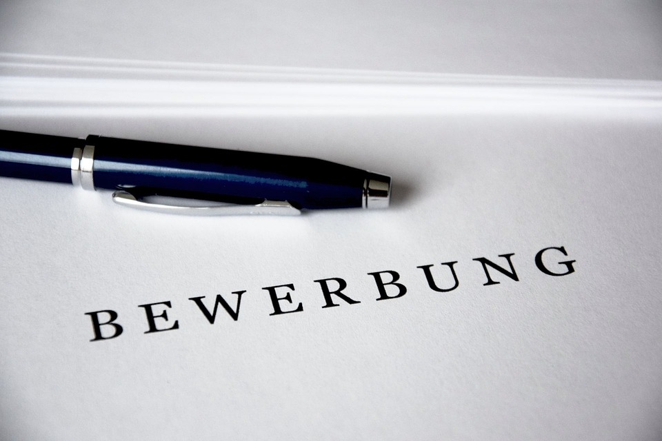 Symbolbild mit schwarzem Bewerbungsschriftzug und einem dunkelblauen Kugelschreiber auf weißem Papierhintergrund.