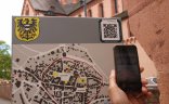 Digitales Gelnhausen Bild eines Stadtplantafel vor der Marienkirche in Gelnhausen. Davor ist eine Hand mit einem Handy das einen QR-Code scannt.