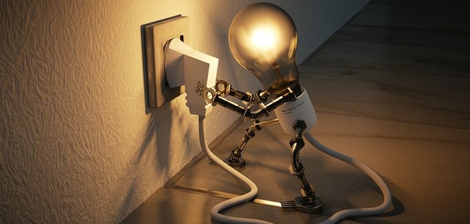 Auf dem Bild ist eine Stilisierte Darstellung einer Glühbirne zu sehne. Diese Glühbirne hat Arme und Beine, sie ist über ein Kabel mit einer Steckdose verbunden durch diese Steckdose bekommt die Glühbirne Strom und leuchtet deshalb. Die Glühbirne hat ihre Arme auf ihrem Stecker und möchte diesen aus der Steckdose ziehen. 