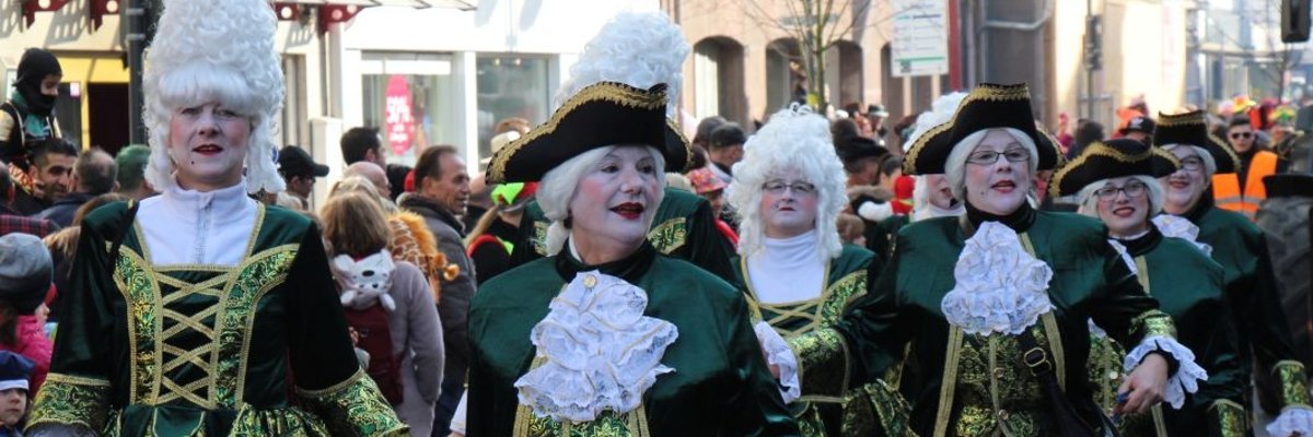 Im Hintergrund ist eine Menschenmenge zu sehen die dem Faschingsumzug zuschaut. Im Vordergrund sieht man mehrere verkleidete Fauen. Die Frauen tragen prunkvolle schwarze Kleider mit grünen und goldenen Verzieherungen. Weiterhin tragen manche der Frauen noch Federhüte.