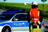 Zu sehen ist ein Feuerwehrmann der mit dem Rücken zur Kamera steht und auf ein Polizeiauto schaut. Der Feuerwehrmann trägt neben seiner typischen Uniform eine orangene Warnweste auf der Feuerwehr steht. 