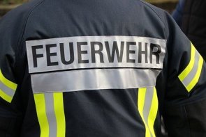 Zu sehen ist eine Nachaufnahme des Rückens eines Feuerwehrmannes. Dieser Trägt seine Uniform, die einem dunkelblauen grundton hat. Auf dieser Uniform sind mehrere Reflektionsstreifen befestigt. Seitlich und auf den Armen sind die Reflektionsstreifen von neongelben Stoffstreifen umgeben. Weiterhin kann man noch den Schriftzug Feuerwehr zentral auf dem Rücken erkennen. 