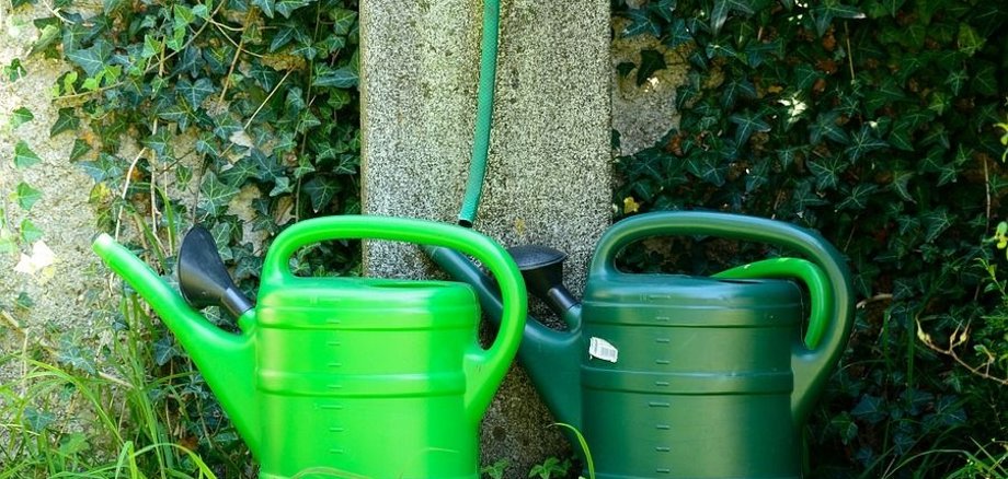 Eine hellgrüne und eine dunkelgrüne Gießkanne mit je 10 Liter Fassungsvermögen stehen an einer Wasserstelle auf einem Friedhof.