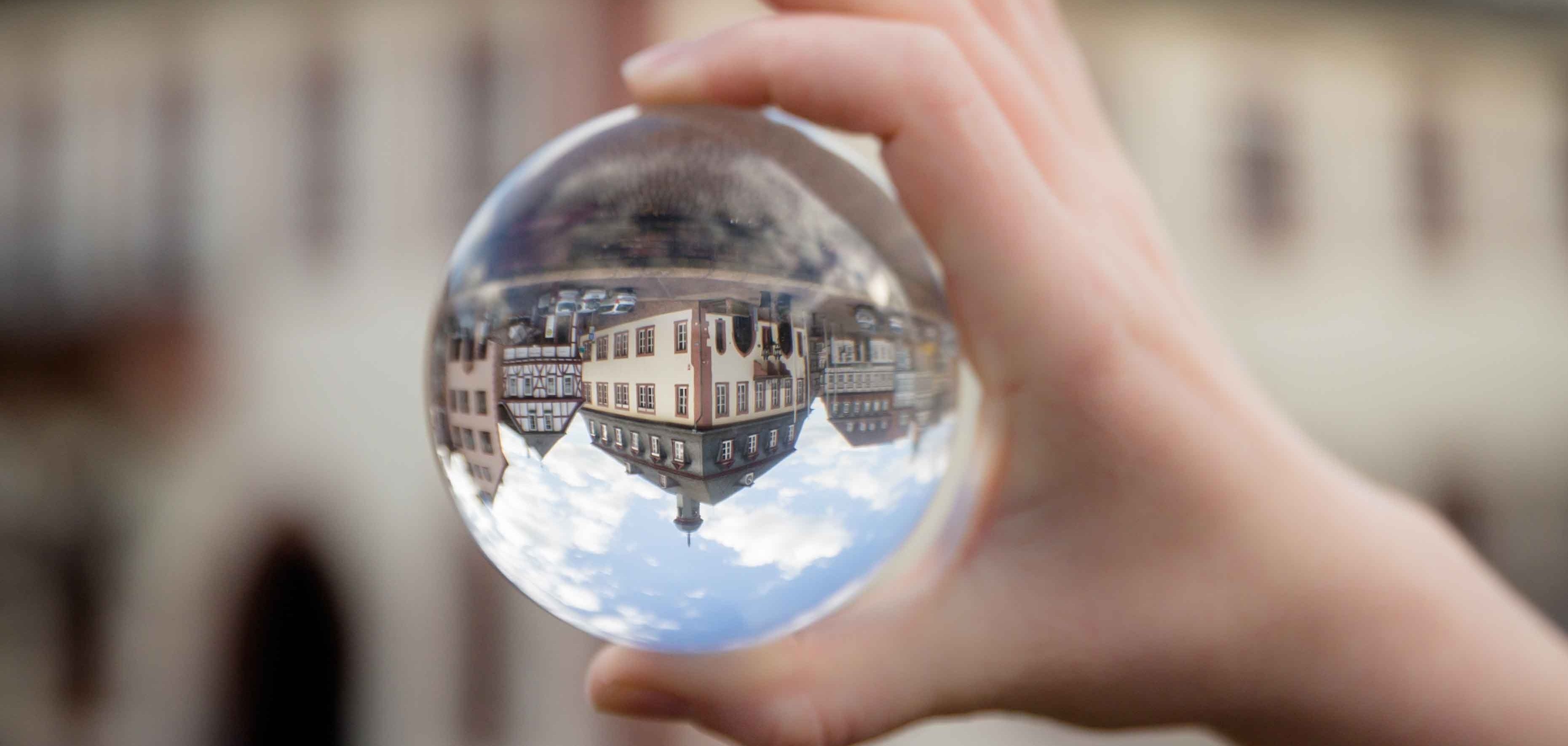 Zu sehen ist eine Hand die eine Glaskugel festhält. In der Glaskugel spiegelt sich verkehrtherum  das Rathaus. 