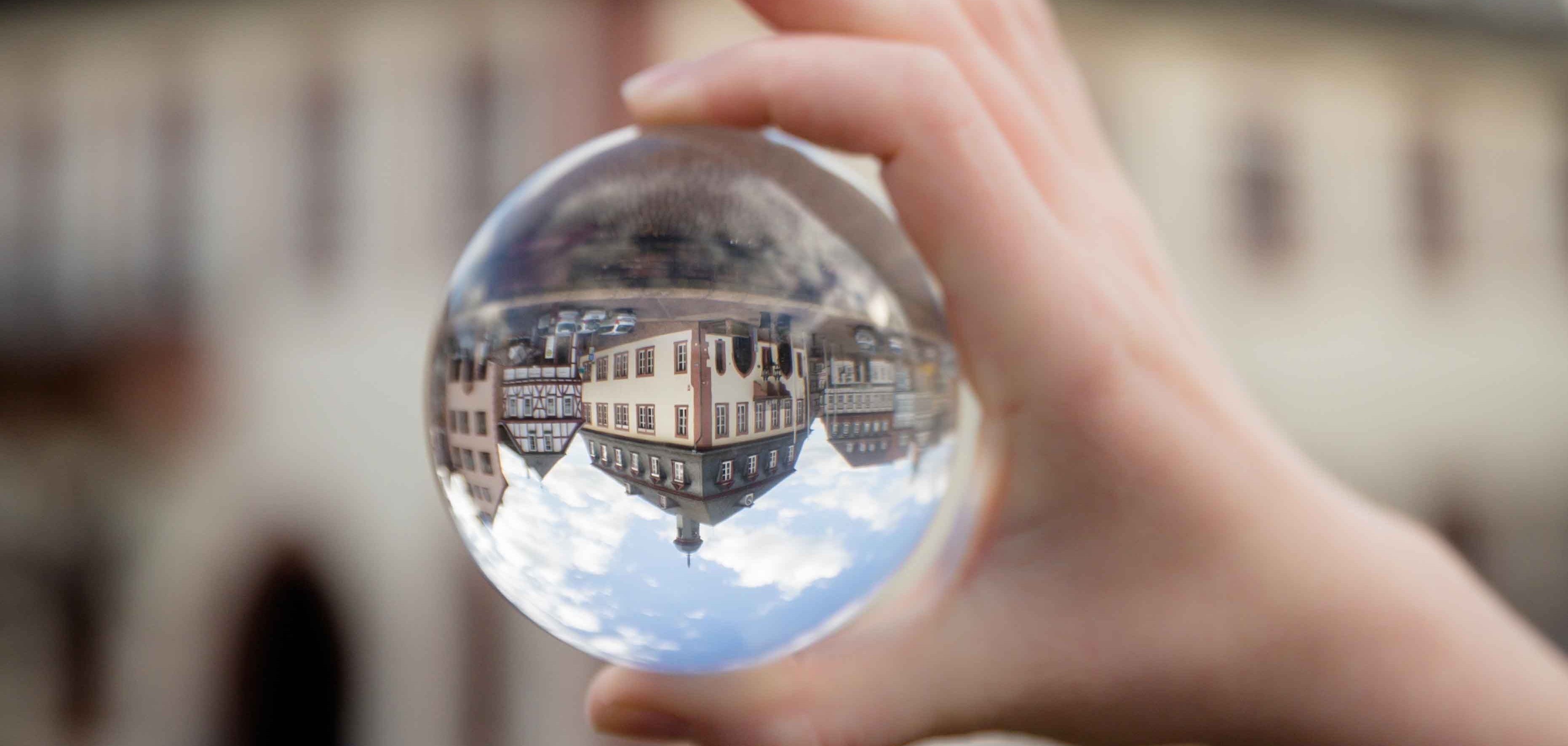 Zu sehen ist eine Hand die eine Glaskugel festhält. In der Glaskugel spiegelt sich verkehrtherum  das Rathaus. 