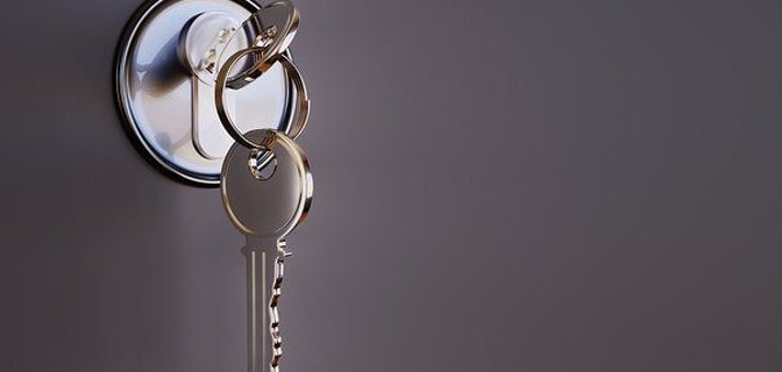 Nahaufnahme eines Türschlosses in einer grauen Tür. In dem runden Türschloss steckt noch ein Schlüssel mit einem Schlüsselring an dem ein weiterer Schlüssel hängt. 